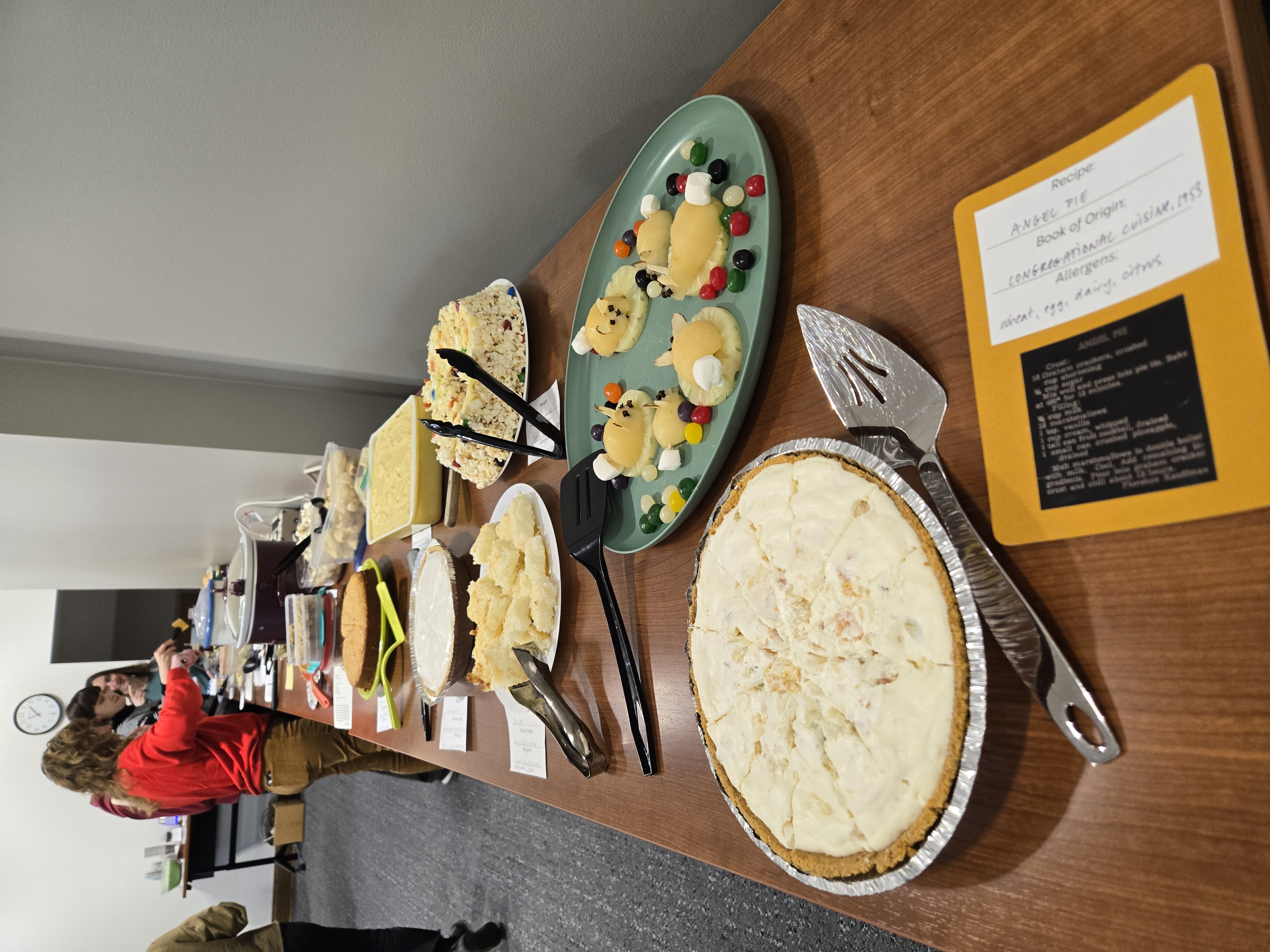 Several people stand with plates at a long table of potluck dishes.