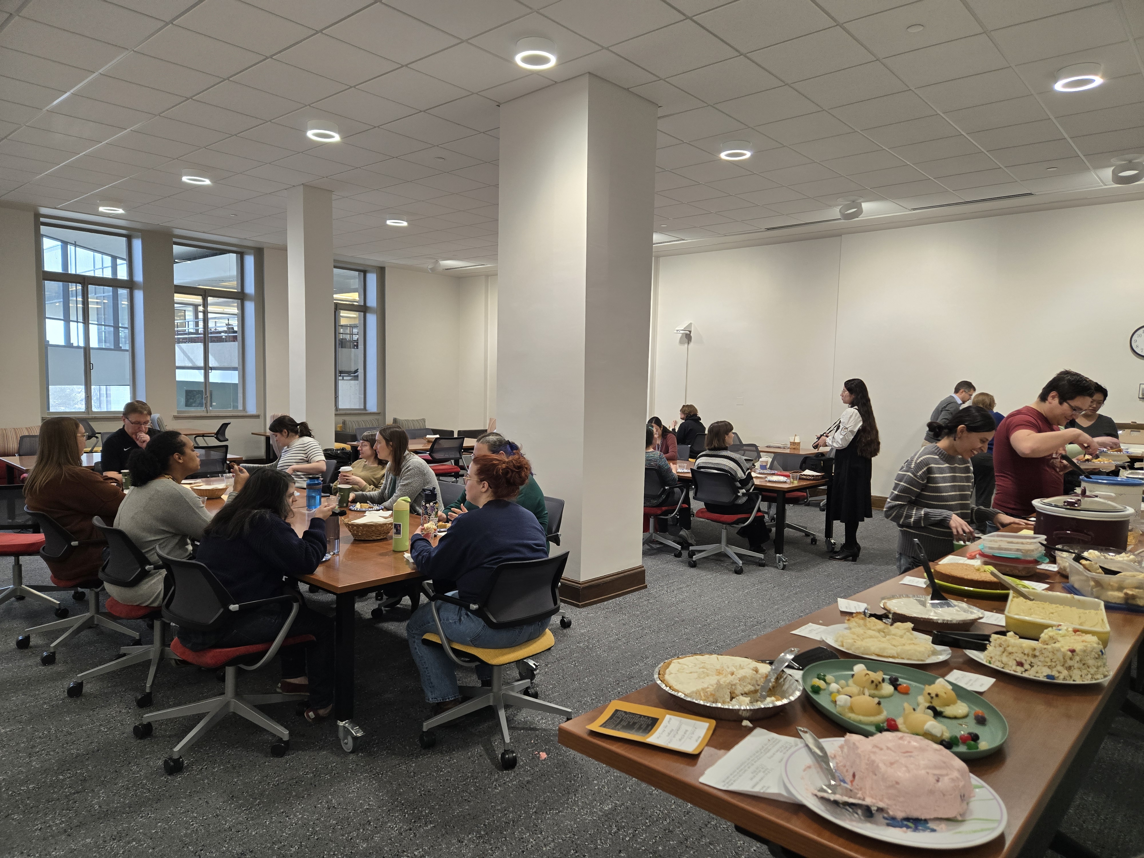 Staff lounge full of people sitting at tables enjoying potluck food.